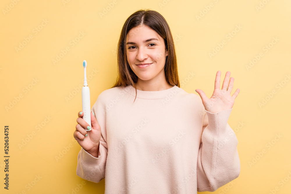 Young caucasian woman holding a electric toothbrush isolated on yellow background smiling cheerful showing number five with fingers. Young caucasian woman holding a electric toothbrush isolated on yellow background smiling cheerful showing number five with fingers.