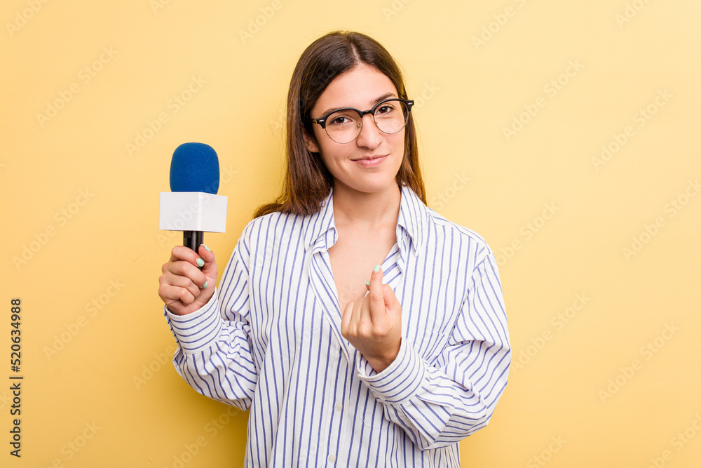 Young caucasian presenter TV woman isolated on yellow background pointing with finger at you as if inviting come closer. Young caucasian presenter TV woman isolated on yellow background pointing with finger at you as if inviting come closer.
