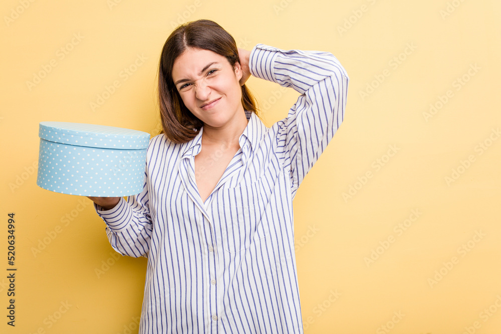 Young caucasian woman holding a gift box isolated on yellow background touching back of head, thinking and making a choice. Young caucasian woman holding a gift box isolated on yellow background touching back of head, thinking and making a choice.