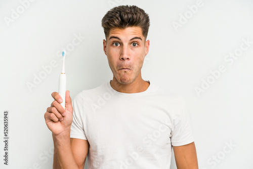 Young caucasian man holding a electric toothbrush isolated on white background shrugs shoulders and open eyes confused.