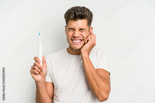 Young caucasian man holding a electric toothbrush isolated on white background covering ears with hands.