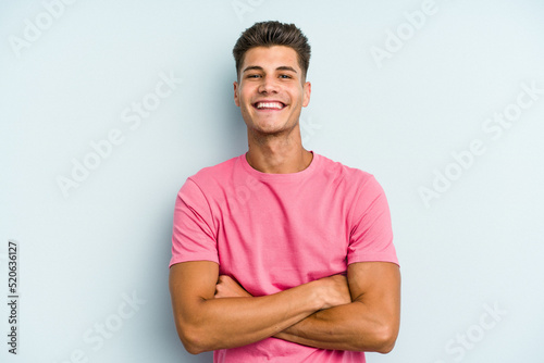 Young caucasian man isolated on blue background who feels confident, crossing arms with determination. © Asier