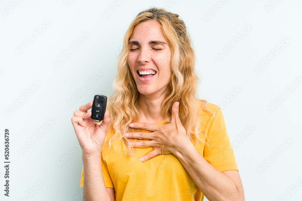 Young caucasian woman holding car keys isolated on blue background laughs out loudly keeping hand on chest. Young caucasian woman holding car keys isolated on blue background laughs out loudly keeping hand on chest.