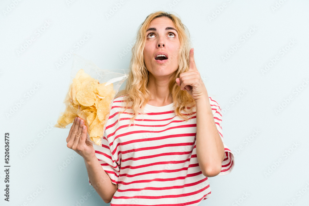 Young caucasian woman holding a bag of chips isolated on blue background pointing upside with opened mouth. Young caucasian woman holding a bag of chips isolated on blue background pointing upside with opened mouth.
