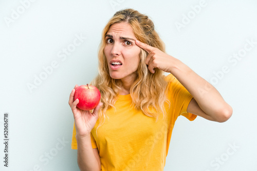 Young caucasian woman holding an apple isolated on blue background showing a disappointment gesture with forefinger.