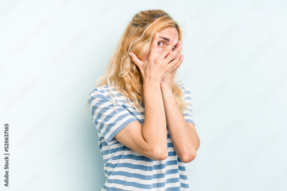 Young caucasian woman isolated on blue background blink through fingers frightened and nervous. Young caucasian woman isolated on blue background blink through fingers frightened and nervous.