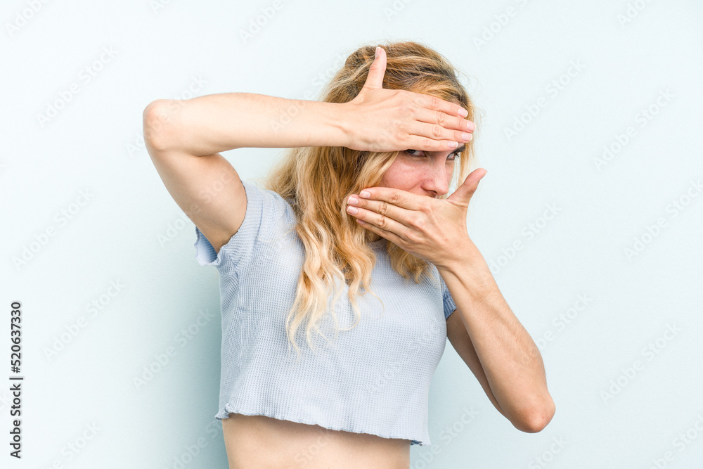 Young caucasian woman isolated on blue background blink at the camera through fingers, embarrassed covering face. Young caucasian woman isolated on blue background blink at the camera through fingers, embarrassed covering face.