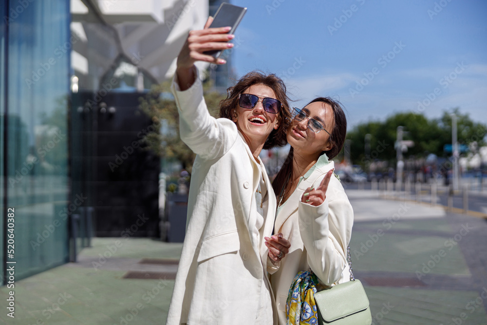 Obraz premium Two smiling woman in trendy outfits making selfie on background of modern building