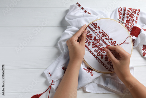 Woman embroidering shirt with red thread at white wooden table, top view and space for text. Ukrainian national clothes photo