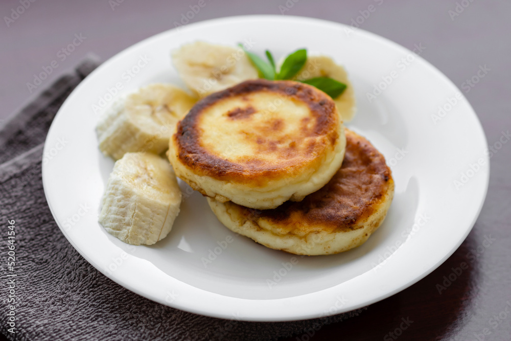 Two cottage cheese pancakes with sliced banana in a white plate. Traditional breakfast or lunch. Traditional Ukrainian breakfast.