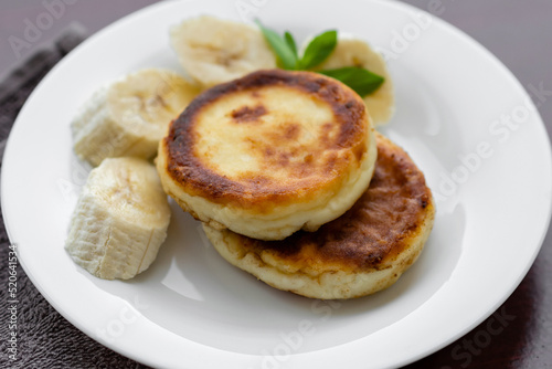 Two cottage cheese pancakes with sliced banana in a white plate. Traditional breakfast or lunch. Traditional Ukrainian breakfast.