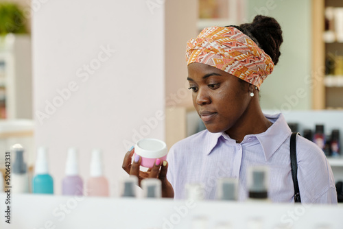 Photography Young black woman holding small pink plastic jar with cream while choosing beaut