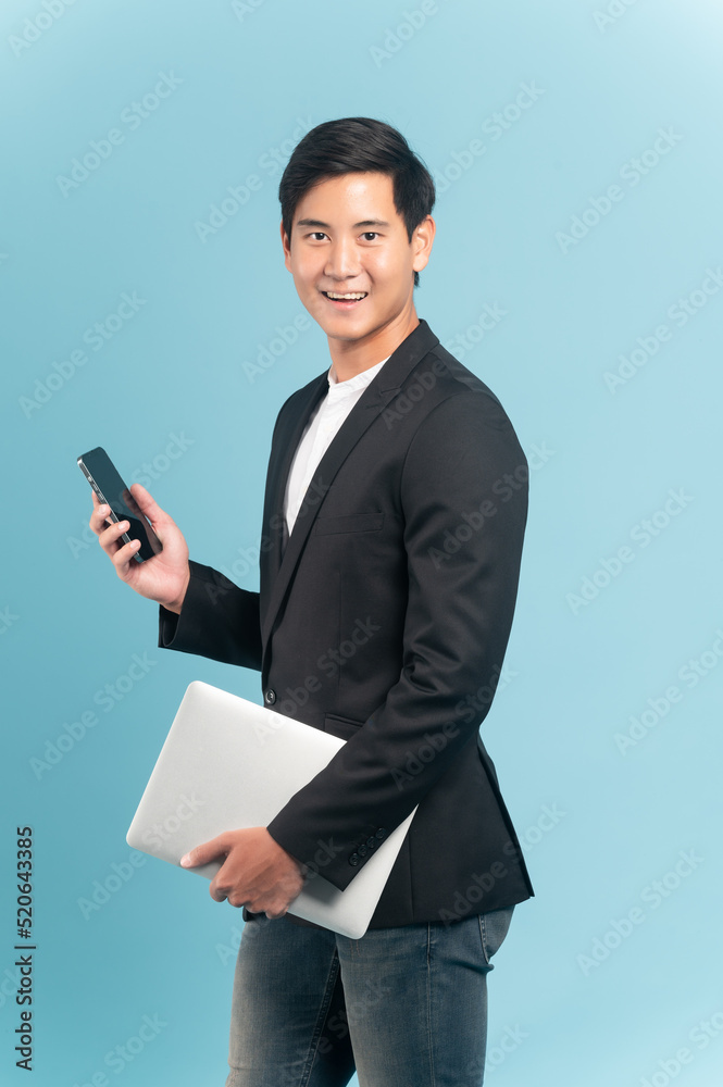 Young asian businessman in smart casual style using smartphone and holding a laptop isolated on blue background Young asian businessman in smart casual style using smartphone and holding a laptop isolated on blue background