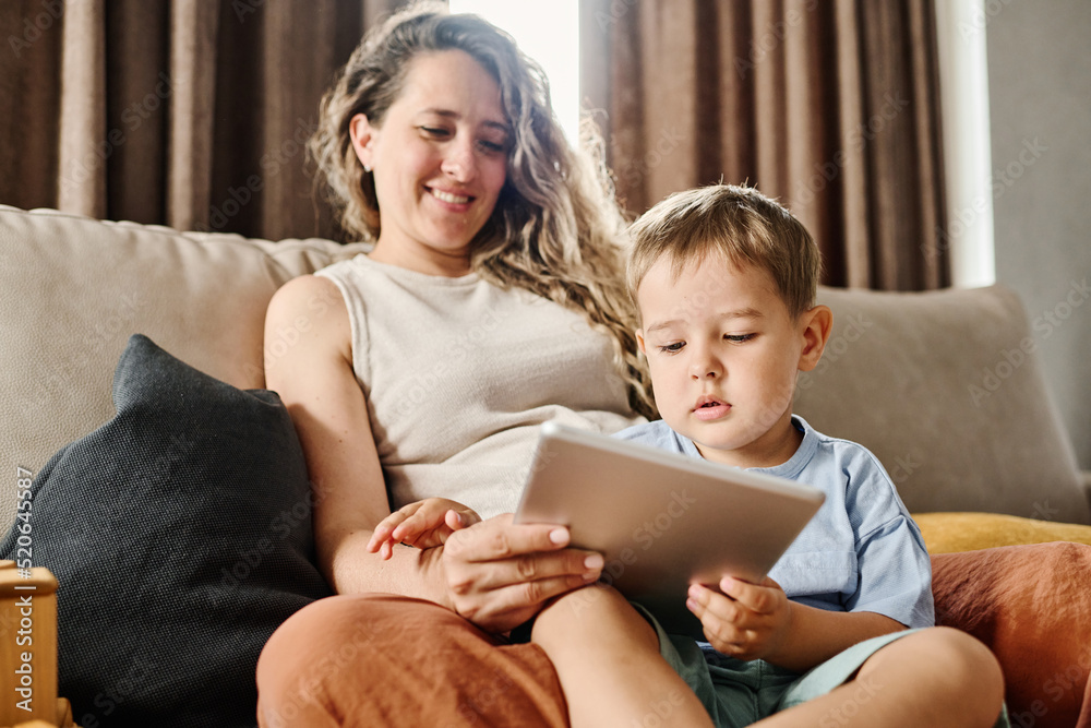 Curious toddler boy looking attentively at tablet screen while watching movie for children or cartoons with his mother at home