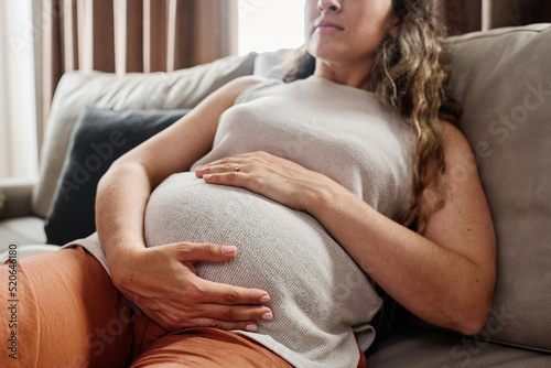 Young pregnant woman keeping hands on her belly while resting on couch at leisure in living room of modern apartment