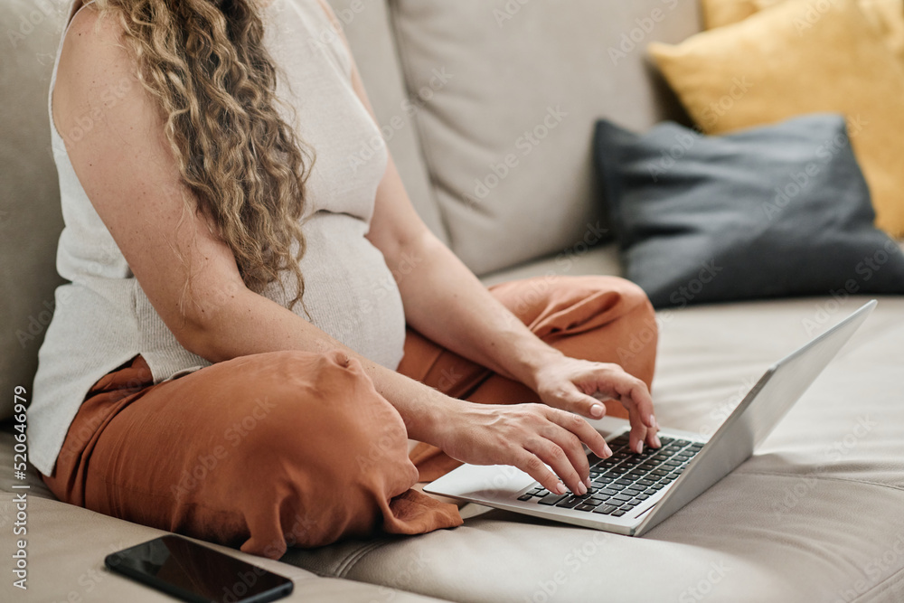 Naklejka premium Cropped shot of young pregnant woman typing on laptop keypad while sitting with crossed legs on couch in living room and networking