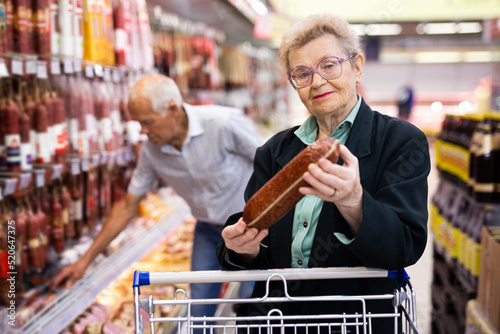 mature woman with glasses picks out salami in the meat section of the supermarket