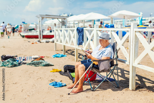 Old woman in a hat sits in a tourist chair on the beach and solves crossword puzzles, a general plan