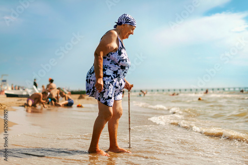 An old woman in a swimsuit with a walking stick comes to the sea on the beach