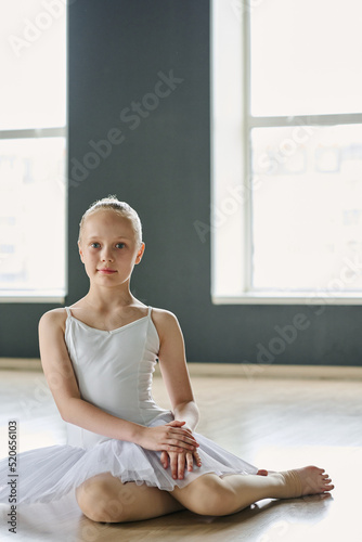 Pretty youthful girl in white tutu and beige tights sitting on the floor of spacious classroom or dance studio and looking at camera