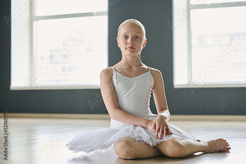 Adolescent schoolgirl in white tutu looking at camera while sitting on the floor of classroom or dance hall for choreography repetitions photo