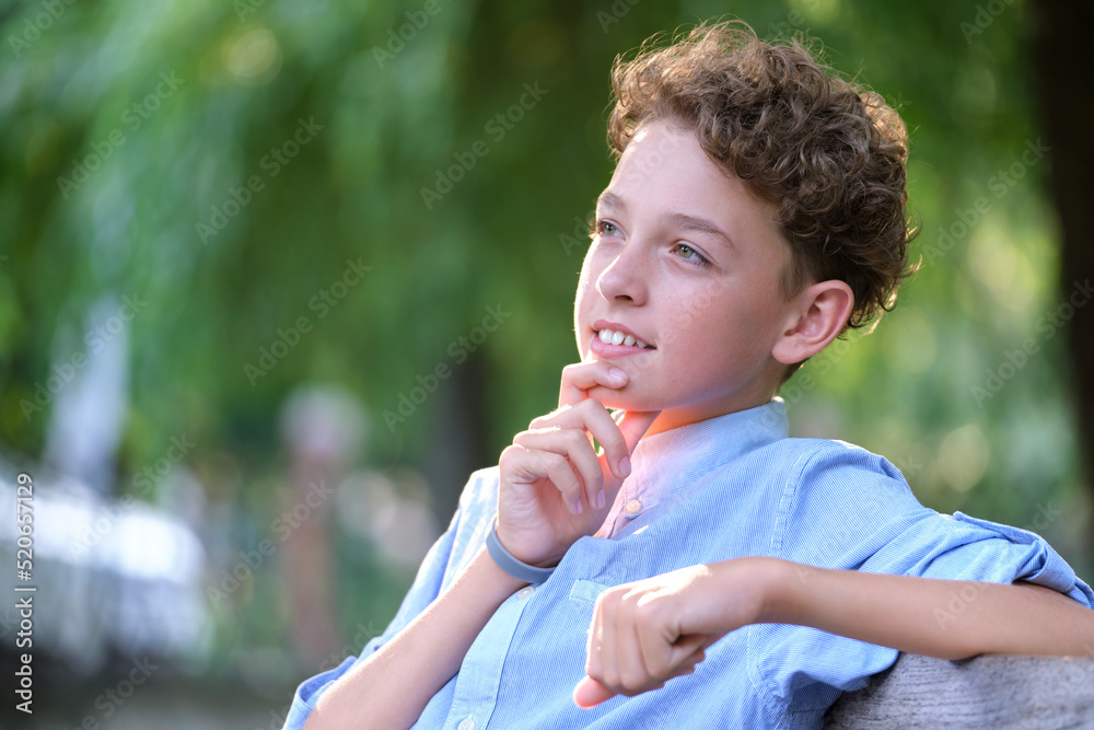 Young happy child boy relaxing sitting on bench in summer park. Positive kid enjoying summertime outdoors. Child wellbeing concept Young happy child boy relaxing sitting on bench in summer park. Positive kid enjoying summertime outdoors. Child wellbeing concept