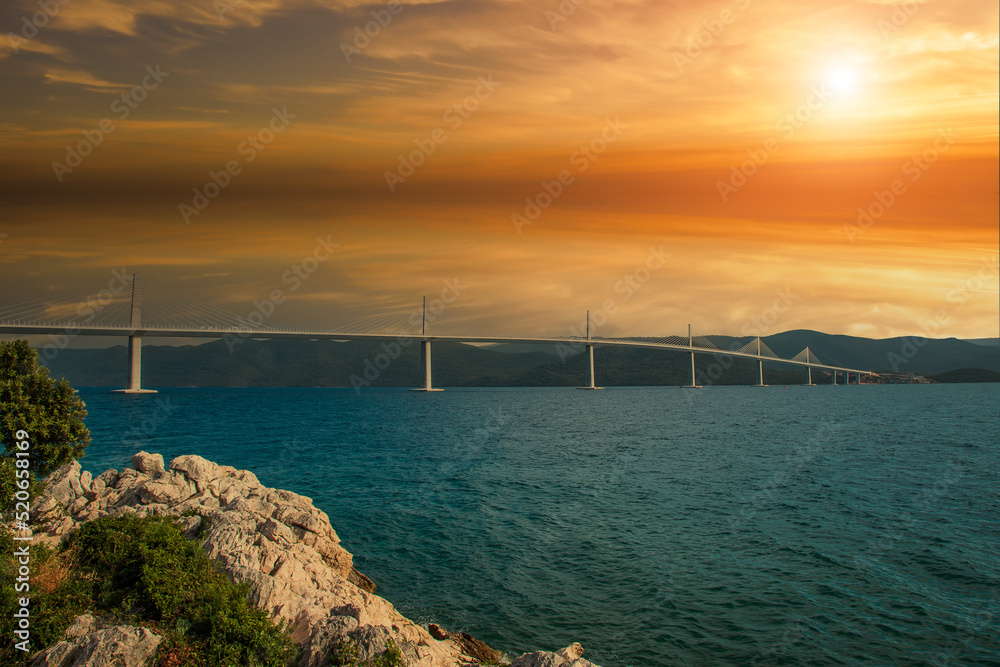 Beautiful modern bridge over the sea on Peljesac in Croatia. In the foreground houses and in the background the Peljesac mountains. Dramatic sky with clouds. Beautiful modern bridge over the sea on Peljesac in Croatia. In the foreground houses and in the background the Peljesac mountains. Dramatic sky with clouds.