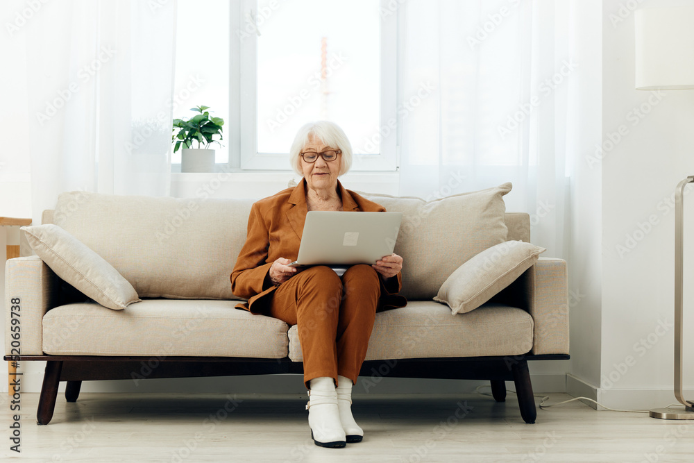 a serious elderly lady is sitting on a beige sofa in a brown suit with a laptop on her lap and enthusiastically working while at home. Full-length photo a serious elderly lady is sitting on a beige sofa in a brown suit with a laptop on her lap and enthusiastically working while at home. Full-length photo