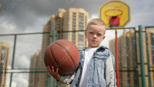 Child boy is standing on basketball court holding ball and looking at camera. © orientka