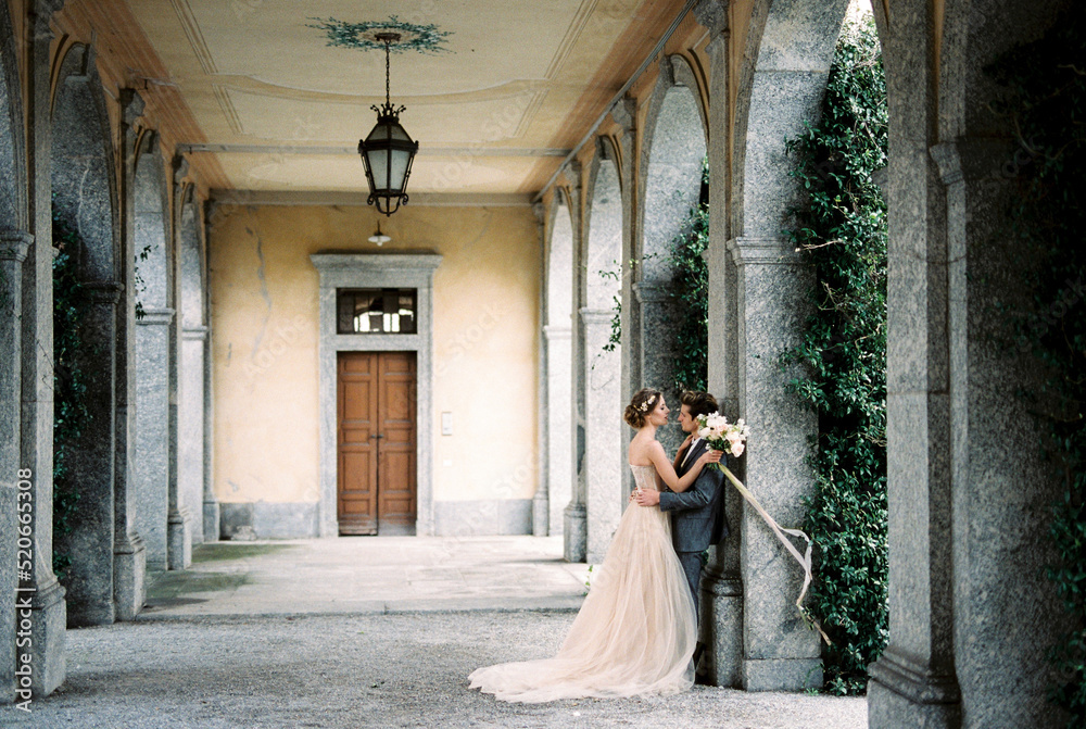 Groom hugs bride leaning against the arch of an old villa. Como, Italy
