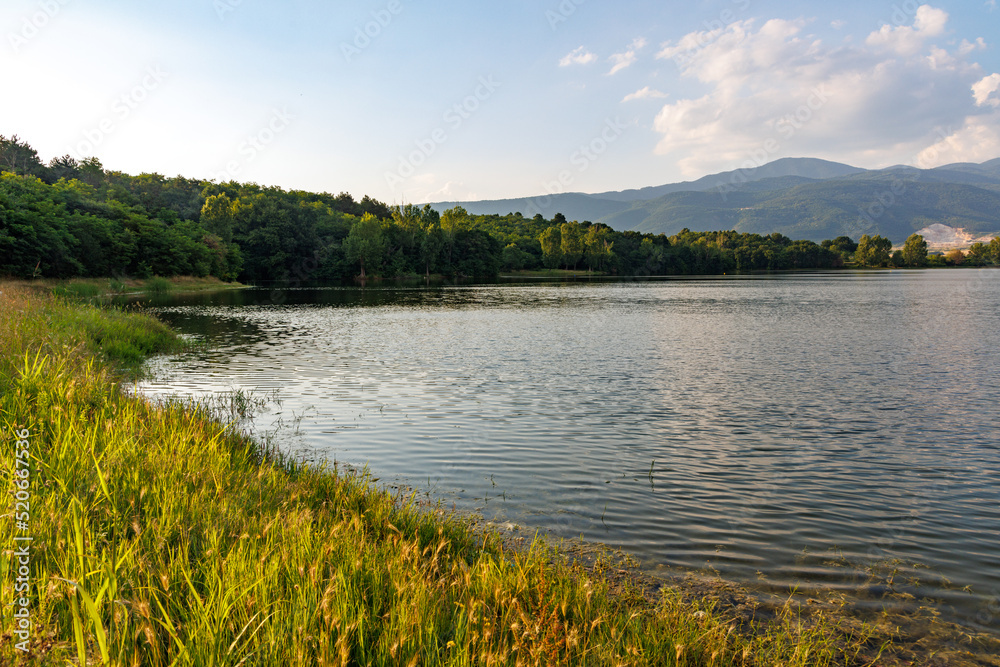 A lake reflects sunlight, against blue sky and mountain ranges and villages