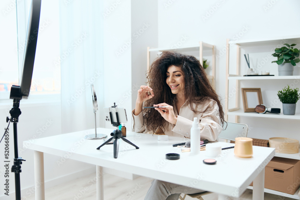 Interested tanned handsome curly Latin beauty blogger in linen shirt tells about brushes in home white interior. Copy space Mockup Banner. Influencer record video review blog using smartphone Interested tanned handsome curly Latin beauty blogger in linen shirt tells about brushes in home white interior. Copy space Mockup Banner. Influencer record video review blog using smartphone