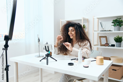 Interested tanned handsome curly Latin beauty blogger in linen shirt tells about brushes in home white interior. Copy space Mockup Banner. Influencer record video review blog using smartphone © SHOTPRIME STUDIO