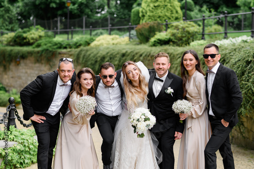 Front view of stylish friends and newlyweds in festival outfits, hugging each other, smiling and posing at camera during wedding walking on park background