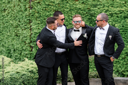 Front view of groom with friends, dressed in black suits and wearing sunglasses, which having fun while posing on background of wall overgrown with plants