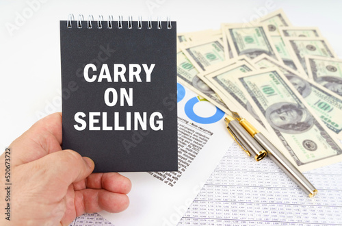 In the man's hand is a notepad with the inscription - Carry on Selling, in the background a pen and dollars