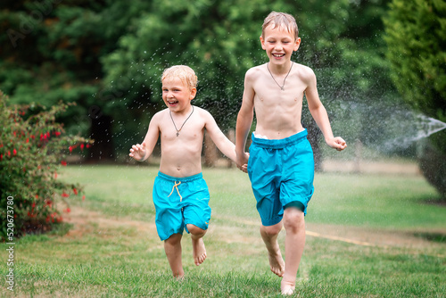 Kids play with water on hot summer day. Children with garden sprinkler. Outdoor fun. Boys run on a field under water drops.