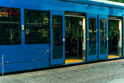 Blue tram with open doors for passengers at a stop on a summer evening, reflections in the windows of the tram, sunlight on the branches of trees outside the windows of the tram, sustainable transport