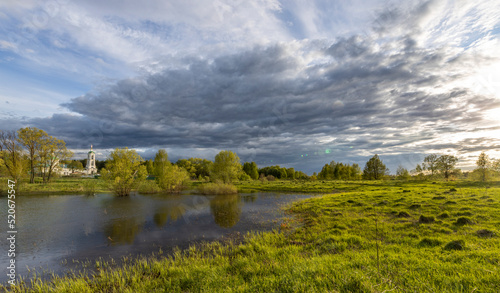 Rural landscape on a spring evening. the dramatic sky is reflected in the river. Bright sun rays illuminate the grass in the foreground.