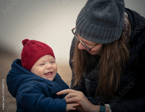 Niño bebé con gorro de lana rojo y campera riendo y mirando a la cámara y jugando con su mamá en la playa en otoño e invierno photo