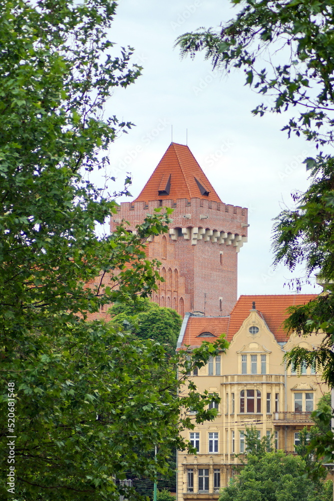 in the photo is a yellow house, trees and a tower of an old red brick castle in the city of Poznan in Poland.
