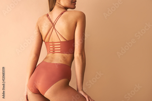 Beautiful, athletic, young woman in underwear poses for the camera with her back turned on beige background.