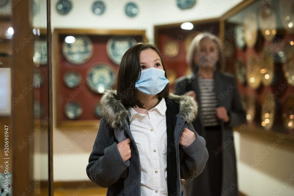 Fototapeta premium Cute interested preteen girl wearing protective face mask exploring artworks in modern museum of applied arts. Forced precautions in pandemic