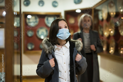 Cute interested preteen girl wearing protective face mask exploring artworks in modern museum of applied arts. Forced precautions in pandemic