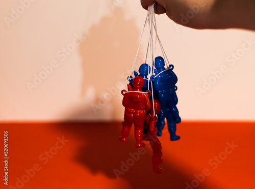 Red and blue Russian soldiers held like puppets by a dictator’s hand on red and white background. Symbol of Russia losing the war.