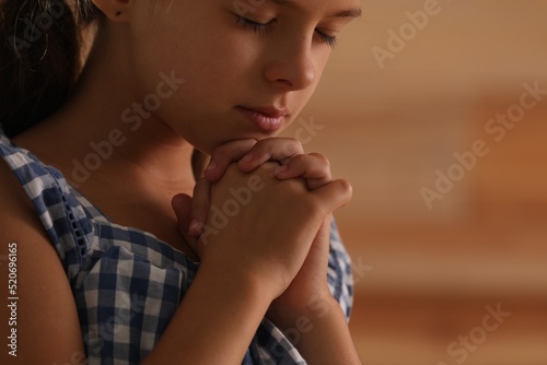 Cute little girl with hands clasped together praying on blurred background, closeup