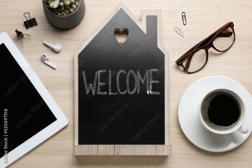 Flat lay composition of house shaped blackboard with word Welcome and gadgets on wooden table Flat lay composition of house shaped blackboard with word Welcome and gadgets on wooden table