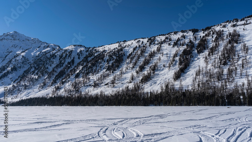 On the snow-covered surface of the frozen lake, traces of tires and snowmobile runners are visible. The forest grows on the mountainside. Clear blue sky. Copy space. Altai. Lower Multinskoe Lake photo