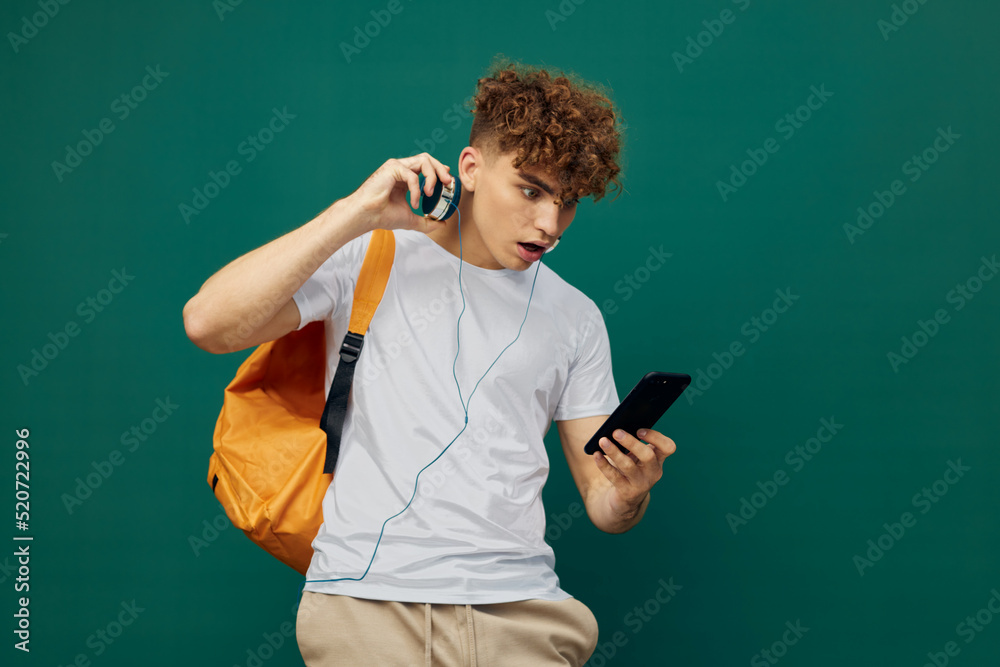 a joyful man, a teenager, stands on a green background listening to music with headphones, holding his smartphone in his hand and looking at it a joyful man, a teenager, stands on a green background listening to music with headphones, holding his smartphone in his hand and looking at it