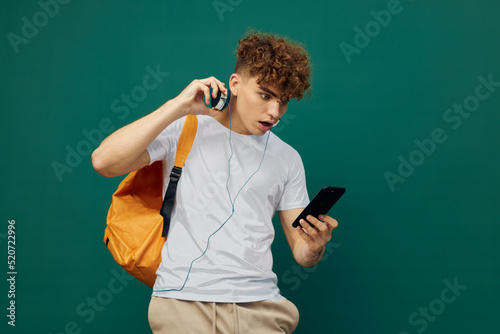 a joyful man, a teenager, stands on a green background listening to music with headphones, holding his smartphone in his hand and looking at it